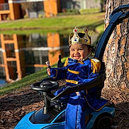 Angie joined the competition — help win amazing prizes! toddler, child, crown, toy_car, blue_outfit, royal_costume, smile, outdoor, tree, pond, grass, sunlight, play, happy, person, vehicle, nature, daytime, costume, portrait