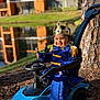 toddler, child, crown, toy_car, blue_outfit, royal_costume, smile, outdoor, tree, pond, grass, sunlight, play, happy, person, vehicle, nature, daytime, costume, portrait
