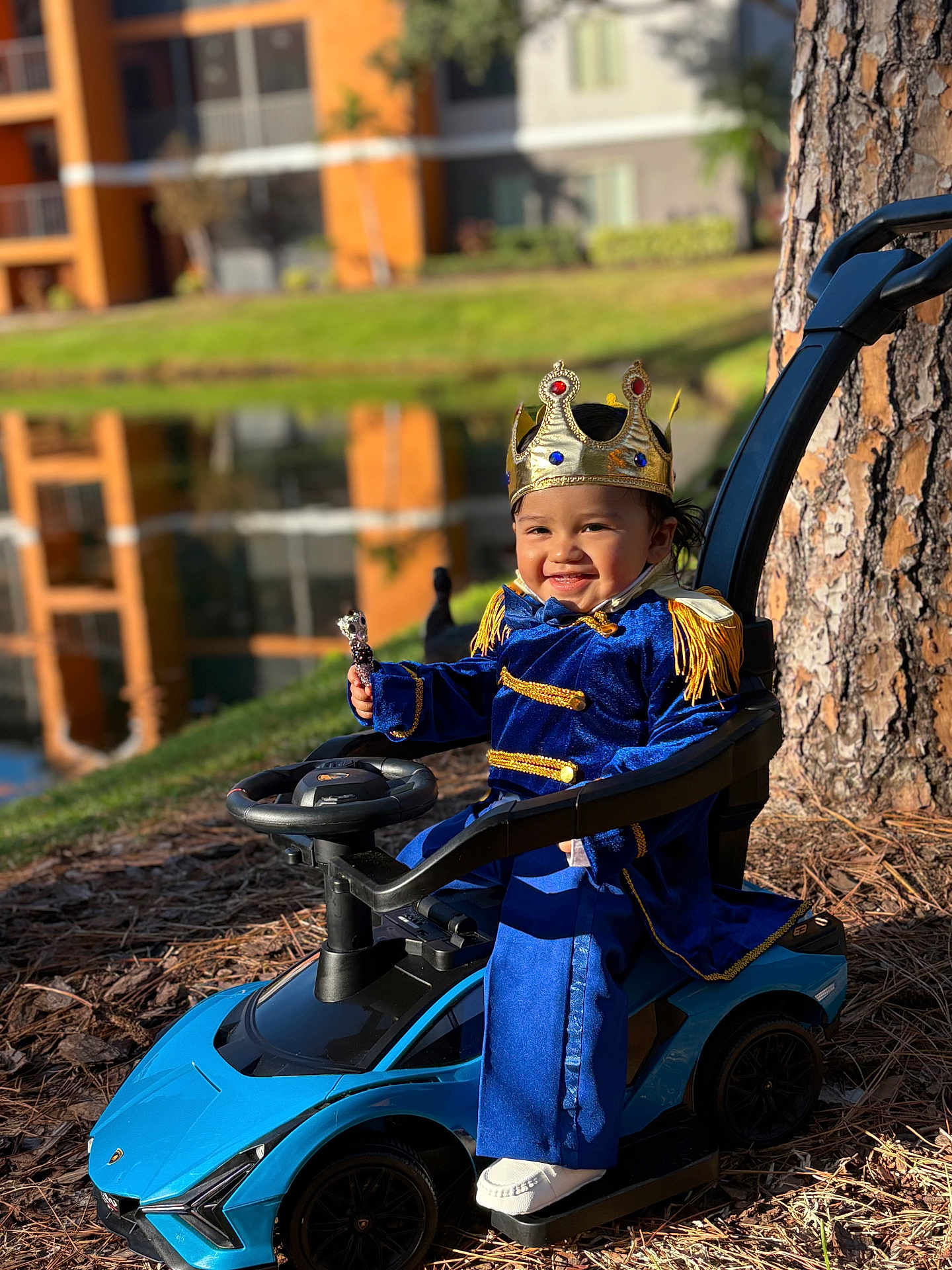 Angie joined the competition — help win amazing prizes! toddler, child, crown, toy_car, blue_outfit, royal_costume, smile, outdoor, tree, pond, grass, sunlight, play, happy, person, vehicle, nature, daytime, costume, portrait