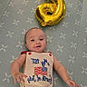 american_flag, baby, balloon, celebration, child, colorful, cute, happy, holiday, indoor, infant, laying_down, mattress, number_3, onesie, patriotic, patterned, person, smiling, toy