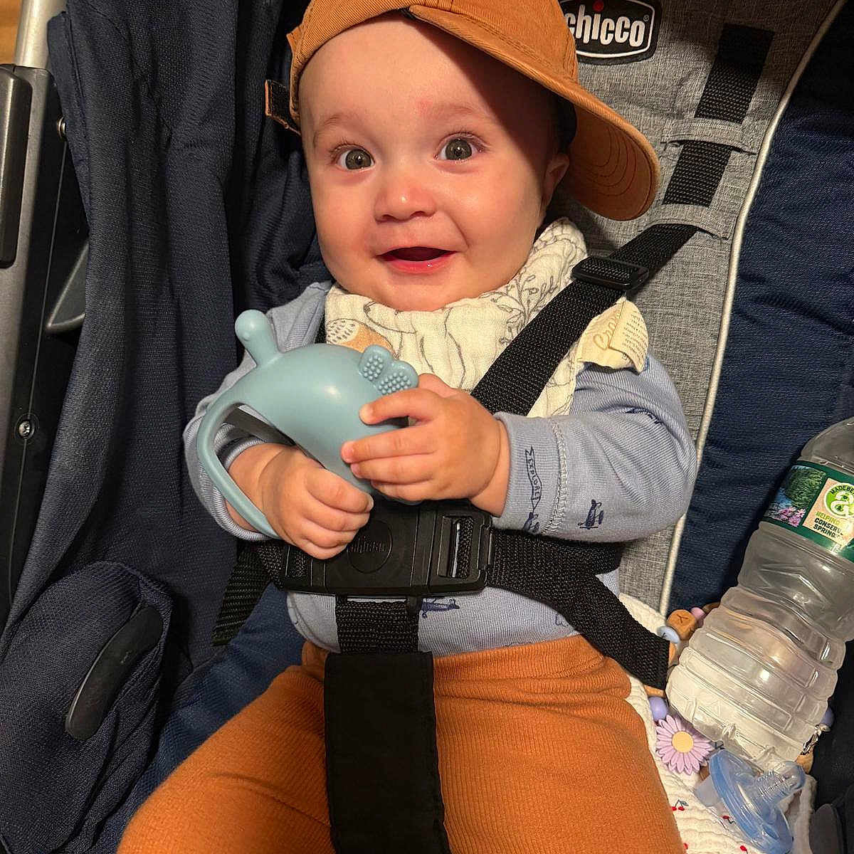Julyan is registered to the contest to win money with this photo: baby, baby_bib, cap, car_seat, child, clothing, face, hand, happy, indoor, infant, orange_pants, pacifier, person, seatbelt, smiling, stroller, teething_toy, toy, water_bottle