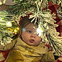 Cody joined the competition — help win amazing prizes! baby, child, face, pine_branch, snow, ornament, gold_ball, christmas_tree, holiday, curious, indoor, red_blanket, yellow_clothing, festive, closeup, decoration, greenery, winter, soft_lighting, cute