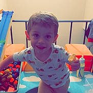 Nickolas is registered to the contest to win money with this photo: baby_bottle, blanket, boy, child, clothing, colorful, floor, furniture, happy, indoor, light, plastic_container, play_area, playtime, railing, shirt, shorts, smile, snack, toy