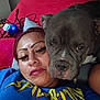 person, dog, pit_bull, selfie, bed, red_blanket, pillow, face, eyes, muzzle, lipstick, eyelashes, lip_piercing, t_shirt, indoor, close_up, pet, companion, chargers, wires