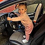 car_interior, car_seat, child, dashboard, daylight, door, floor_mat, hair_buns, hand_grip, orange_wall, pink_clothing, seat_fabric, serious_expression, shoes, side_mirror, standing, steering_wheel, toddler, vehicle, window