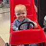 Kamden is registered to the contest to win money with this photo: baby, child, smiling, toy_car, red, striped_onesie, indoor, shopping_mall, floor, wheels, steering_wheel, person, clothing, cute, happy, seat, head, footwear, face, playful