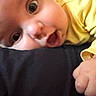 baby, close_up, face, hand, yellow_clothing, curious_expression, infant, skin, person, child, cute, wide_eyes, mouth_open, resting, indoor, soft_light, human, portrait, small_hand, adorable