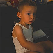 Léo a rejoint le concours — aidez-le/la à gagner de superbes lots ! child, boy, indoor, sitting, white_tank_top, shorts, toys, couch, wooden_wall, room, warm_lighting, face, expression, furniture, casual, home, curious, looking, young, person