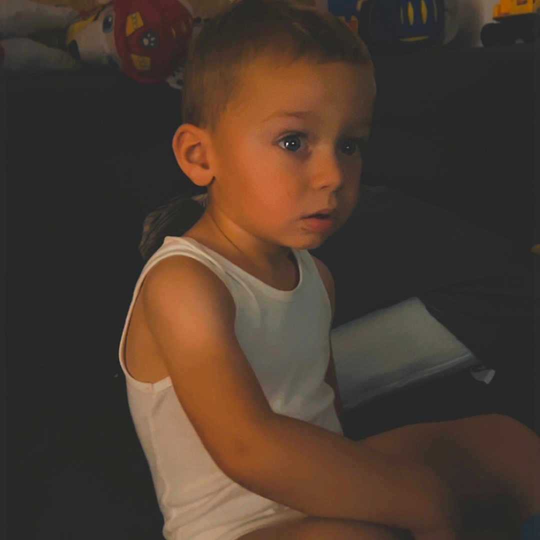Léo a rejoint le concours — aidez-le/la à gagner de superbes lots ! boy, casual, child, couch, curious, expression, face, furniture, home, indoor, looking, person, room, shorts, sitting, toys, warm_lighting, white_tank_top, wooden_wall, young