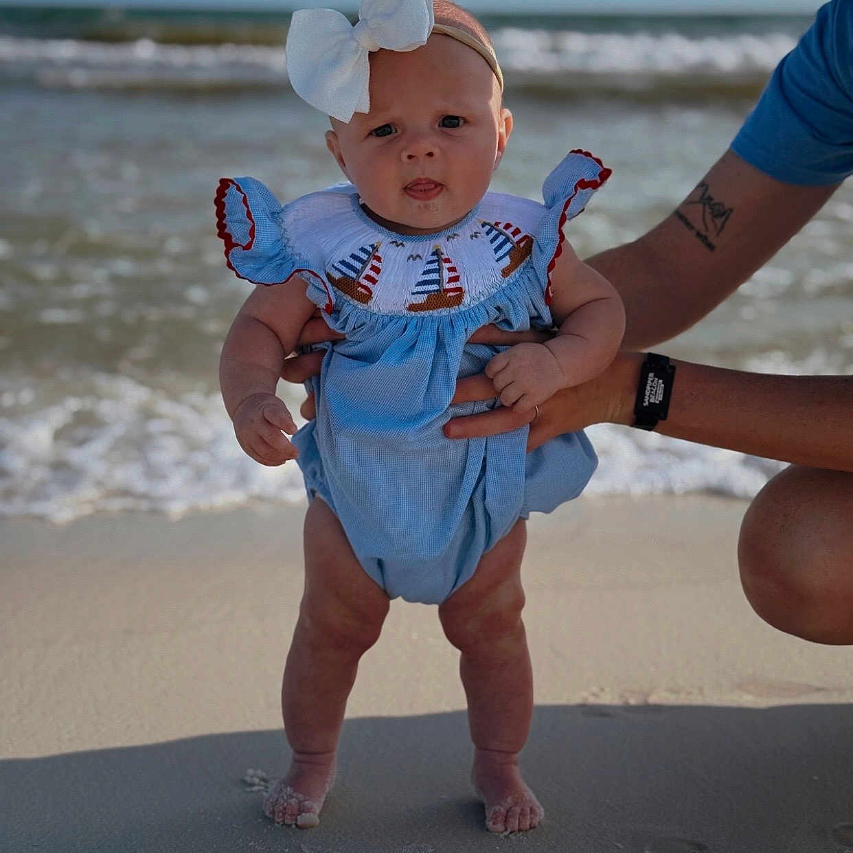 Eleanor joined the competition — help win amazing prizes! baby, child, beach, sand, ocean, water, waves, blue_clothing, headband, bow, person, infant, summer, sunlight, outdoor, hands, tattoo, watch, footprints, cute