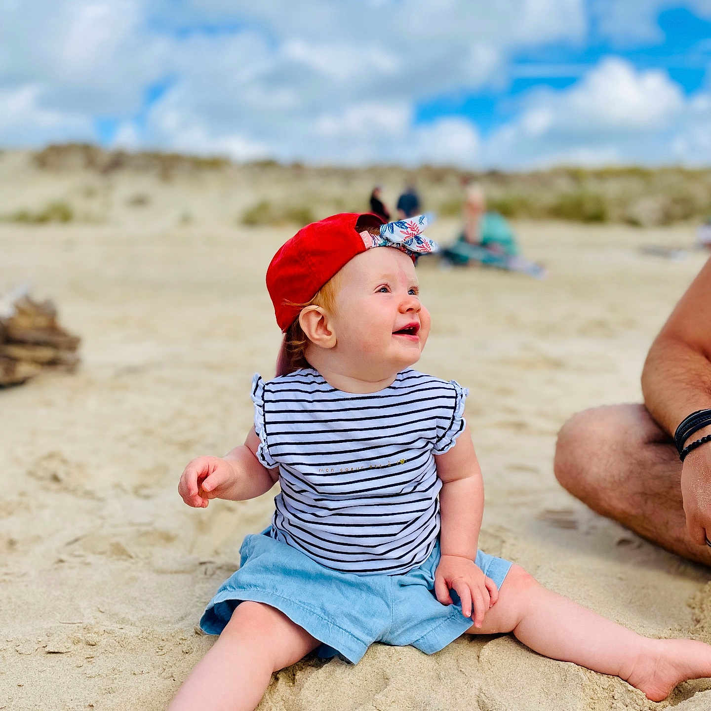 Thessa participe au concours pour gagner de l'argent avec cette photo : adult_arm, baby, barefoot, beach, blue_shorts, casual, child, clouds, happy, nature, outdoor, person, red_cap, sand, sitting, sky, smiling, striped_shirt, summer, sunny