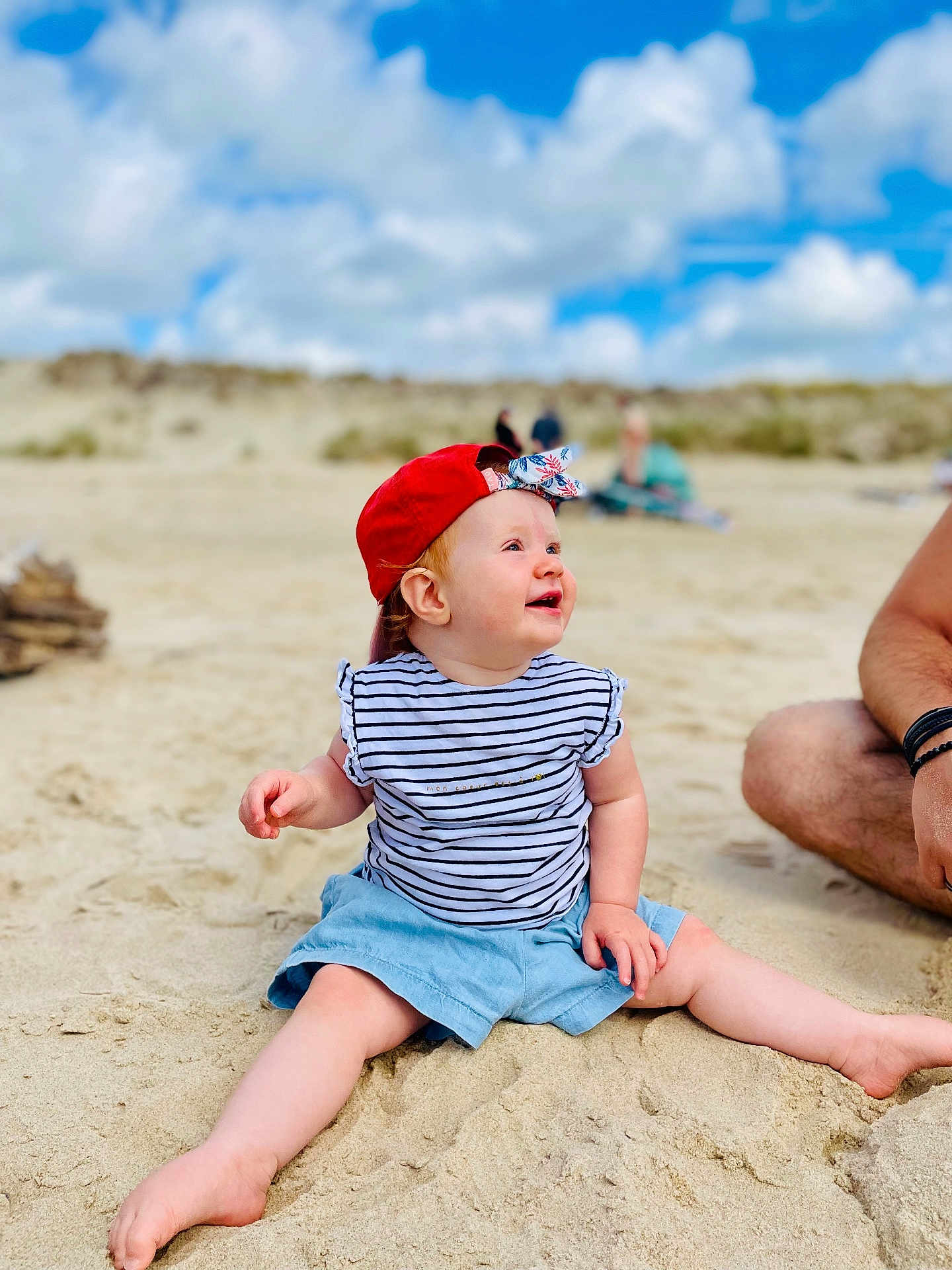 Thessa participe au concours pour gagner de l'argent avec cette photo : baby, child, beach, sand, sky, clouds, red_cap, striped_shirt, blue_shorts, barefoot, smiling, sitting, person, adult_arm, outdoor, sunny, nature, happy, summer, casual