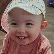 John is registered to the contest to win money with this photo: app_icons, cap, casual_wear, child, clothing, dolphin_pattern, face, floor, furniture, hands, happy, home, indoor, living_room, pet_accessory, smile, table, toddler, weather_widget, wooden_floor