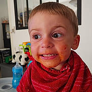 Mattéo participe au concours pour gagner de l'argent avec cette photo : toddler, child, messy_face, red_bib, high_chair, indoor, playful_expression, food_stains, short_hair, big_eyes, home, toy, table, furniture, person, cute, smiling, face, clothing, baby