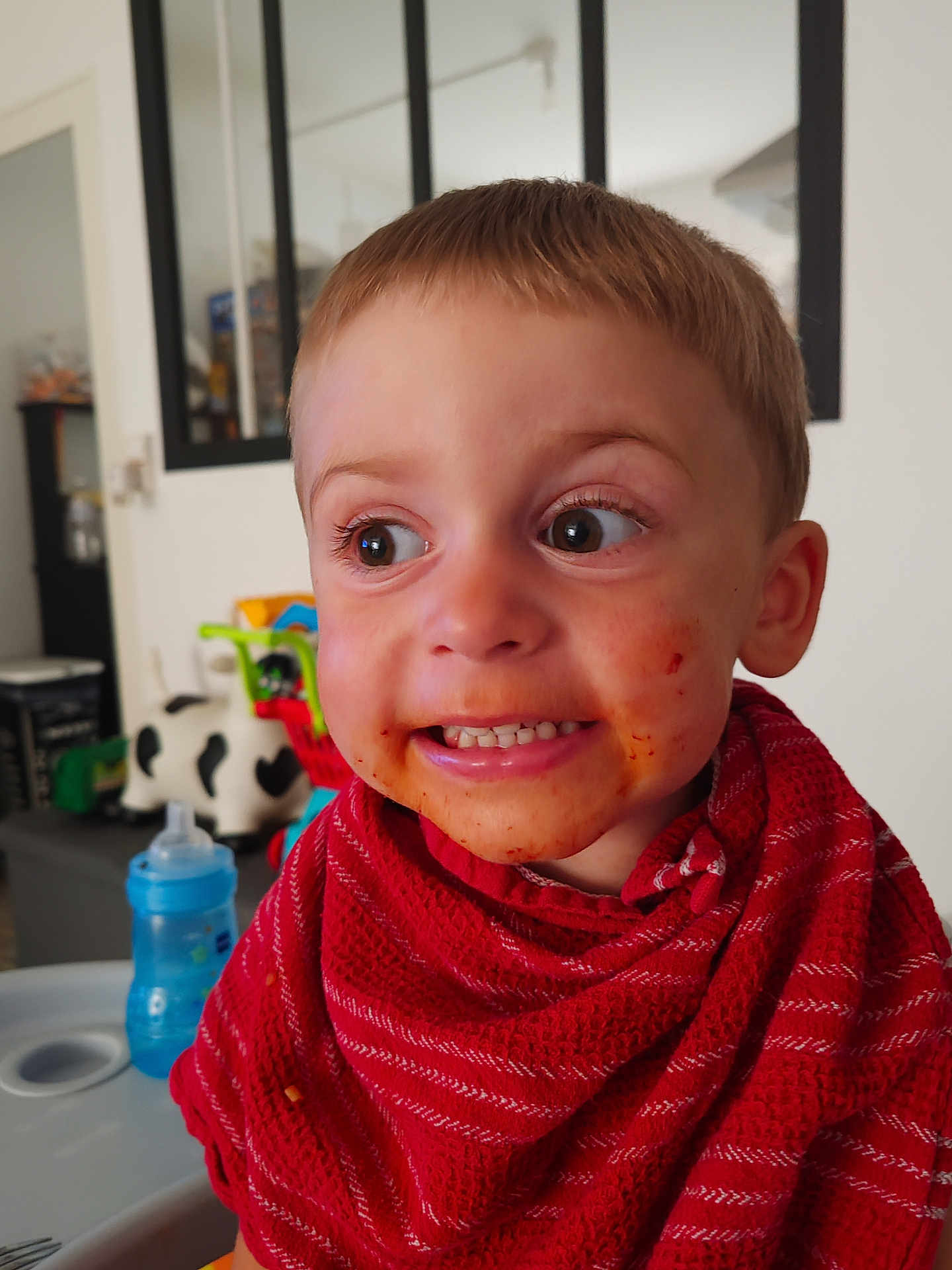 Mattéo participe au concours pour gagner de l'argent avec cette photo : toddler, child, messy_face, red_bib, high_chair, indoor, playful_expression, food_stains, short_hair, big_eyes, home, toy, table, furniture, person, cute, smiling, face, clothing, baby