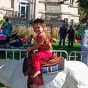 Mason is registered to the contest to win money with this photo: child, pony, horse, saddle, barefoot, outdoor, park, grass, people, building, daytime, smiling, red_pants, brown_shirt, fence, flowers, trees, sunlight, crowd, event