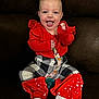 baby, smiling, sitting, red_clothing, patterned_pants, pacifier, pacifier_clip, happy, infant, indoor, couch, furniture, playful, tongue_out, cute, child, person, cozy, bright_colors, portrait