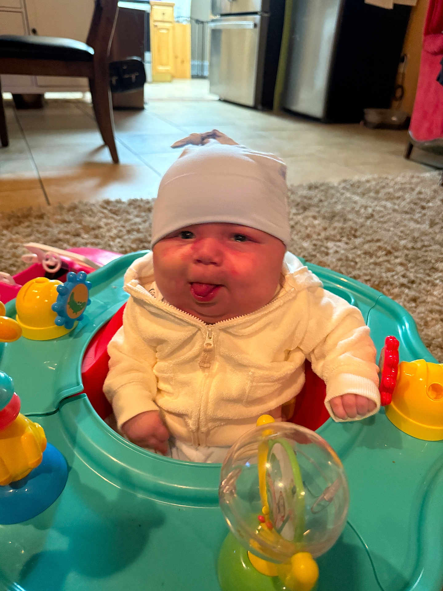 Jaxson joined the competition — help win amazing prizes! baby, child, tongue_out, white_hoodie, hat, indoor, activity_center, toys, colorful, carpet, kitchen, floor, furniture, playful, infant, person, cute, smiling, home, seated