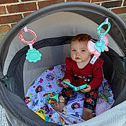 Kiana is registered to the contest to win money with this photo: baby, baby_girl, blanket, blue_eyes, brick_wall, child, colorful, curious, cute, face, floral_pants, indoor, infant, mesh_sides, playful, playpen, red_clothing, sitting, soft_toys, toys