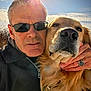 man, dog, golden_retriever, selfie, outdoor, sunlight, sunglasses, portrait, face, hand, ring, fur, pet, animal, closeup, nature, sky, casual, warm_light, affection