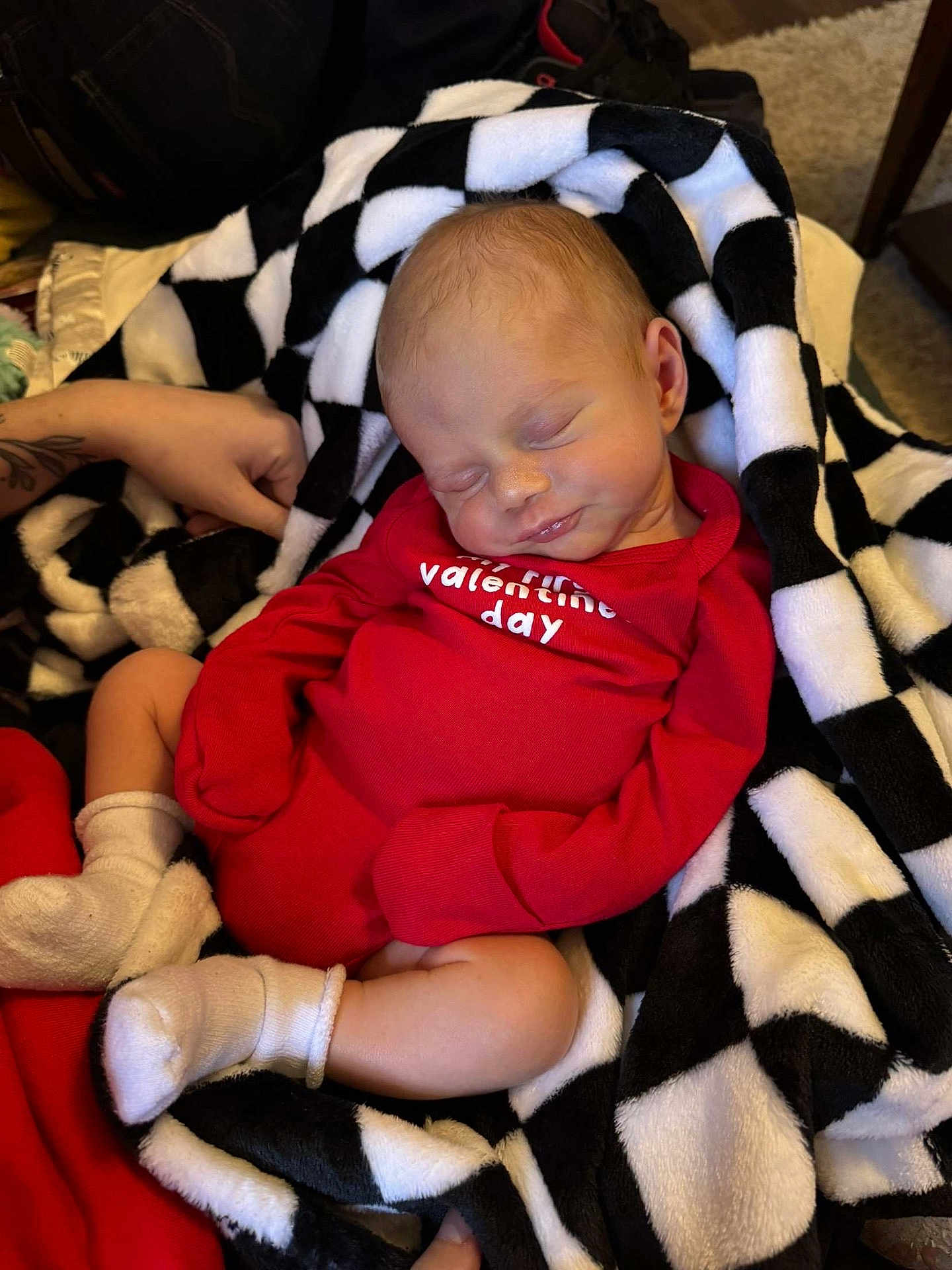 Kash joined the competition — help win amazing prizes! baby, newborn, sleeping, blanket, red_clothing, onesie, white_socks, tattoo, hand, indoor, cozy, comfort, peaceful, infant, cute, soft, rest, black_and_white, checkered_pattern, portrait