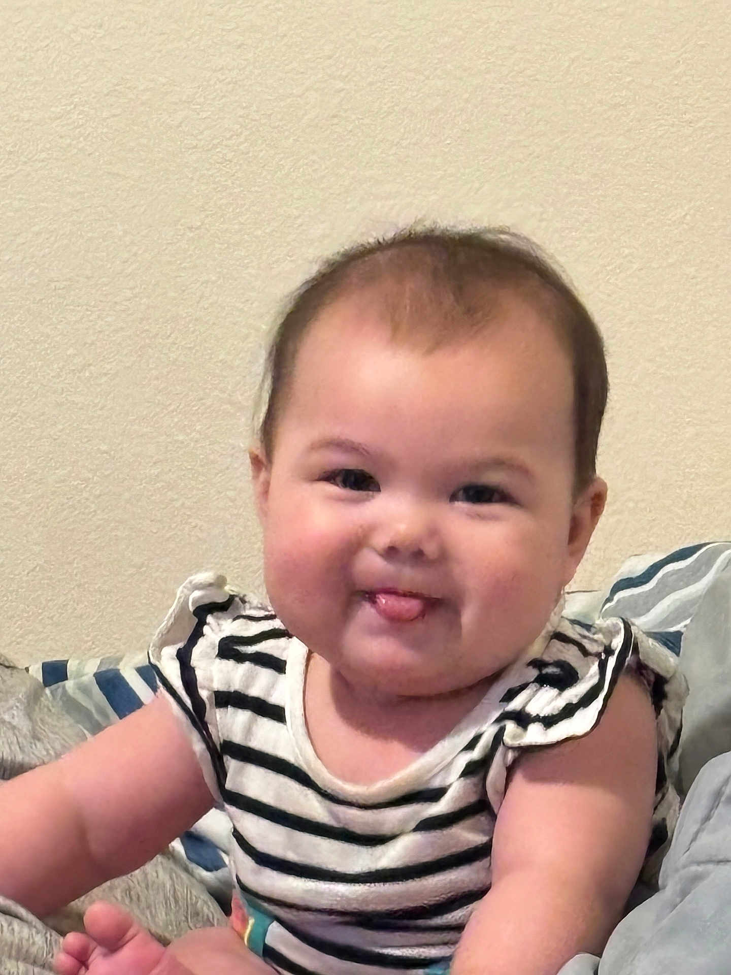 Harlee is registered to the contest to win money with this photo: baby, child, face, tongue, striped_shirt, arm, pillow, blanket, smile, cheeks, infant, sitting, cute, indoors, person, young_child, skin, hair, happy, portrait