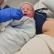 Emerald is registered to the contest to win money with this photo: newborn, baby, hospital, swaddled, blanket, hat, gloved_hand, person, tattoo, arm, bed, medical, resting, infant, sleeping, closeup, healthcare, patient, care, indoors
