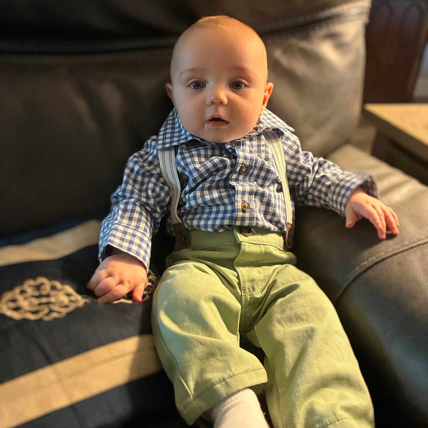 Liam joined the competition — help win amazing prizes! baby, child, infant, sitting, couch, leather, shirt, checkered_shirt, suspenders, pants, green_pants, socks, white_socks, face, eyes, hand, indoor, furniture, portrait, cute