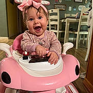 Madison is registered to the contest to win money with this photo: baby, chair, child, clothing, dining_room, face, footwear, hands, happy, headwear, home, indoor, person, pink_bow, pink_toy_car, smile, table, toy, walker, wooden_floor