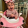 baby, chair, child, clothing, dining_room, face, footwear, hands, happy, headwear, home, indoor, person, pink_bow, pink_toy_car, smile, table, toy, walker, wooden_floor