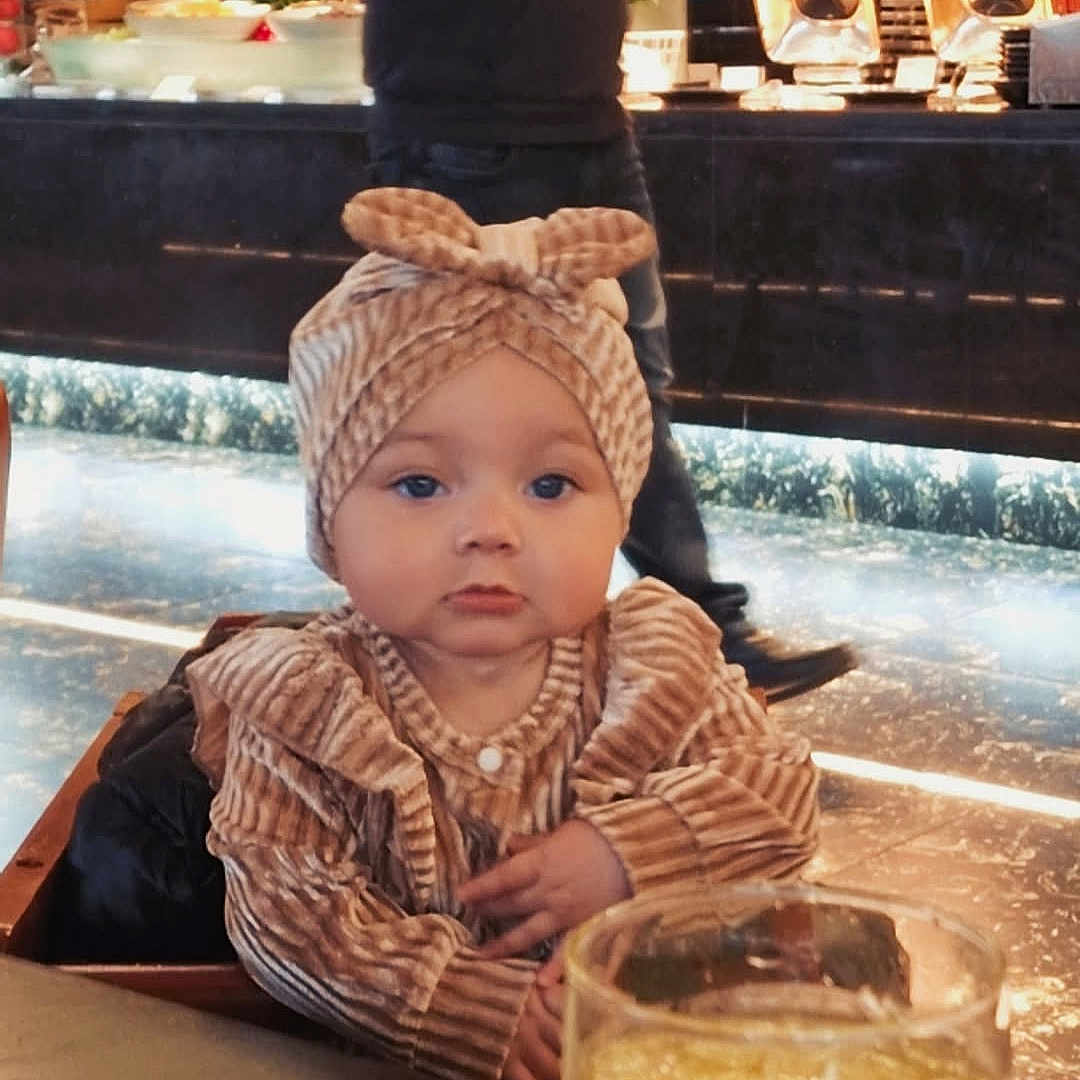 Shaniyah joined the competition — help win amazing prizes! adult, baby, blurred_background, casual_clothing, child, cozy, cute, drink, food, glass, headband, indoor, infant, orange_juice, person, portrait, restaurant, seated, striped_clothing, table