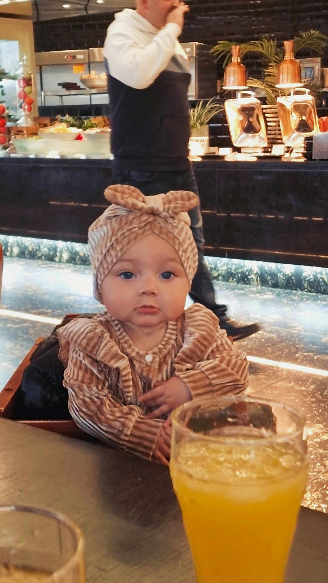 Shaniyah joined the competition — help win amazing prizes! baby, child, infant, headband, striped_clothing, table, glass, orange_juice, drink, indoor, person, adult, blurred_background, restaurant, food, casual_clothing, seated, cute, portrait, cozy