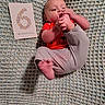 Auryn is registered to the contest to win money with this photo: baby, infant, child, six_months_old, lying_down, smiling, face, hands, feet, red_shirt, gray_pants, knitted_blanket, milestone_card, indoor, portrait, close_up, sitting, playful, family, cozy