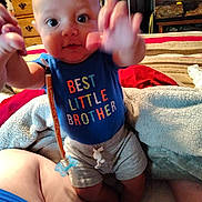 Auryn is registered to the contest to win money with this photo: baby, child, infant, sitting, smiling, wide_eyes, portrait, indoors, bedroom, blanket, cushion, clothing, blue_shirt, text_on_clothing, best_little_brother, raised_hands, reaching, toy, pacifier, furniture