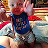 baby, child, infant, sitting, smiling, wide_eyes, portrait, indoors, bedroom, blanket, cushion, clothing, blue_shirt, text_on_clothing, best_little_brother, raised_hands, reaching, toy, pacifier, furniture