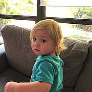 Louis is registered to the contest to win money with this photo: toddler, child, blonde_hair, blue_shirt, couch, window, indoor, portrait, young_child, seated, looking_back, natural_light, casual_clothing, home_interior, curtains, greenery_outside, expression, face, person, furniture
