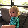 toddler, child, blonde_hair, blue_shirt, couch, window, indoor, portrait, young_child, seated, looking_back, natural_light, casual_clothing, home_interior, curtains, greenery_outside, expression, face, person, furniture