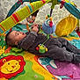 baby, play_mat, colorful, toy, plush_toy, child, infant, indoor, curtain, pattern, lion, elephant, clothing, gray_outfit, smiling, lying_down, hand, play_gym, fabric, cute