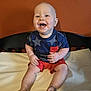 Xaiden is registered to the contest to win money with this photo: baby, child, smiling, crib, blue_shirt, red_shorts, happy, sitting, indoor, toddler, face, person, clothing, furniture, head, skin, expression, joyful, cute, background
