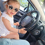 Karima a rejoint le concours — aidez-le/la à gagner de superbes lots ! child, toddler, sunglasses, car, steering_wheel, white_dress, bracelet, person, window, trees, interior, seat, gear_shift, sunlight, dashboard, reflection, side_mirror, adult, casual_clothing, seatbelt