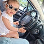 child, toddler, sunglasses, car, steering_wheel, white_dress, bracelet, person, window, trees, interior, seat, gear_shift, sunlight, dashboard, reflection, side_mirror, adult, casual_clothing, seatbelt