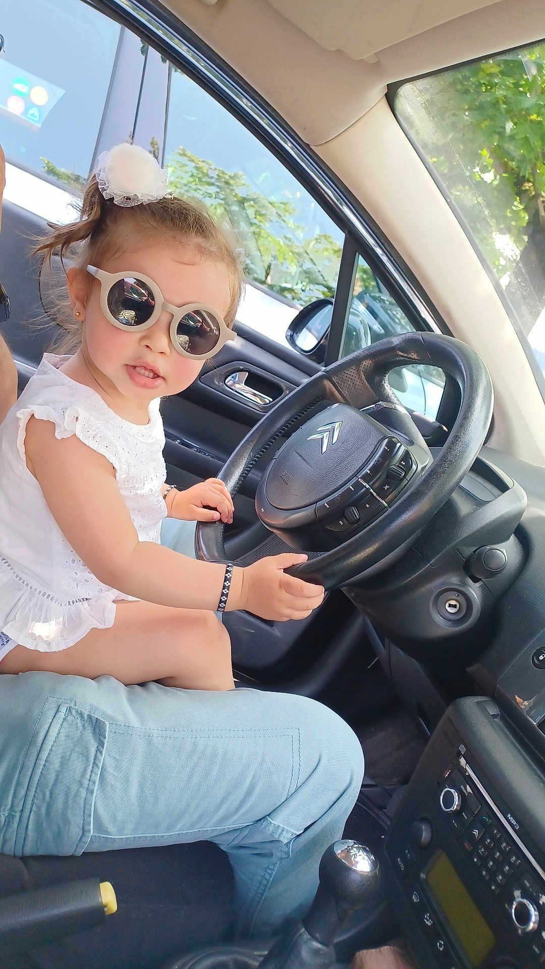 Karima a rejoint le concours — aidez-le/la à gagner de superbes lots ! child, toddler, sunglasses, car, steering_wheel, white_dress, bracelet, person, window, trees, interior, seat, gear_shift, sunlight, dashboard, reflection, side_mirror, adult, casual_clothing, seatbelt