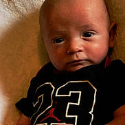 Holden is registered to the contest to win money with this photo: baby, infant, child, face, eyes, hand, clothing, black_shirt, number_23, logo, blanket, lying_down, portrait, cute, skin, head, expression, young, indoors, person
