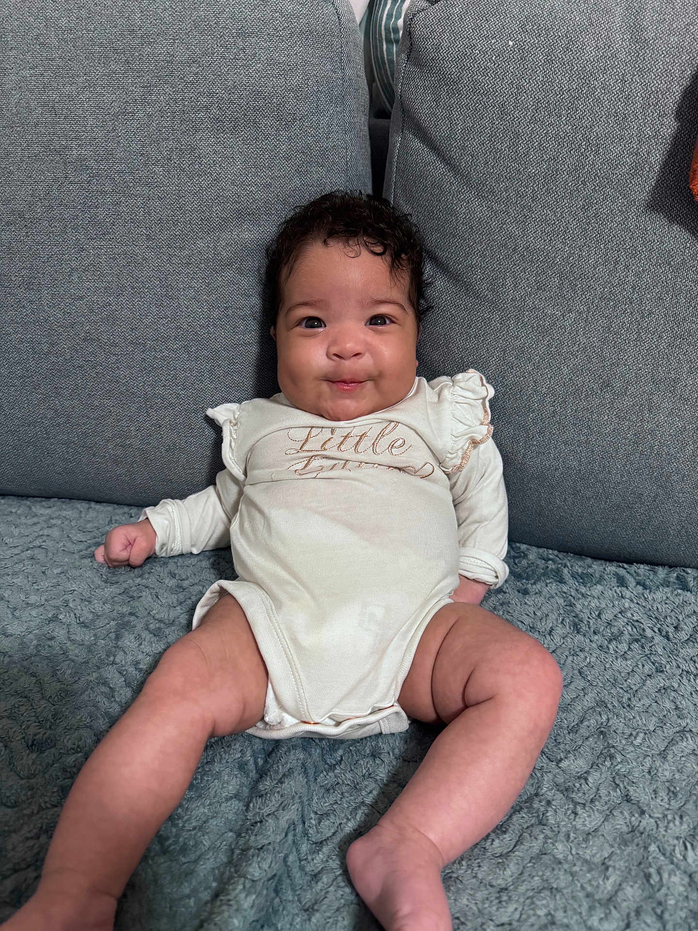 Olivia is registered to the contest to win money with this photo: baby, infant, smile, curly_hair, onesie, couch, blanket, chubby_cheeks, cute, sitting, indoors, child, person, happy, portrait, young, skin, comfortable, cozy, expression