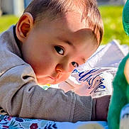 Ezio participe au concours pour gagner de l'argent avec cette photo : baby, child, infant, face, skin, hair, clothing, sleeve, blanket, pattern, toy, plush, green, outdoor, grass, daylight, portrait, curious, lying_down, closeup