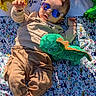 baby, child, sunglasses, balloon, plush_toy, dinosaur_toy, blanket, pattern, elephant_pattern, clothing, pants, socks, outdoor, sunlight, shadow, cute, casual, celebration, relaxing, portrait