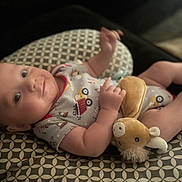 Dylan is registered to the contest to win money with this photo: baby, infant, smiling, plush_toy, cushion, patterned, clothing, indoor, cute, lying_down, child, person, soft_toy, relaxed, happy, small_hands, head, face, eyes, cozy