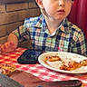 child, boy, pizza, food, table, checkered_tablecloth, plaid_shirt, cap, phone, plate, slice, messy_face, restaurant, brick_wall, red_booth, cutting_board, utensil, casual_clothing, indoor, curious_expression