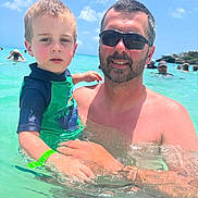 Lorenzo is registered to the contest to win money with this photo: child, man, water, beach, ocean, swimming, sunlight, sunglasses, sky, clouds, wristband, green_shirt, holding, outdoor, vacation, summer, people, family, happy, waves