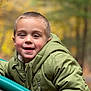 child, boy, smile, face, portrait, green_jacket, jacket, outdoor, autumn, bokeh, close_up, happy, playground, park, short_hair, eyes, hood, zipper, cheeks, soft_lighting
